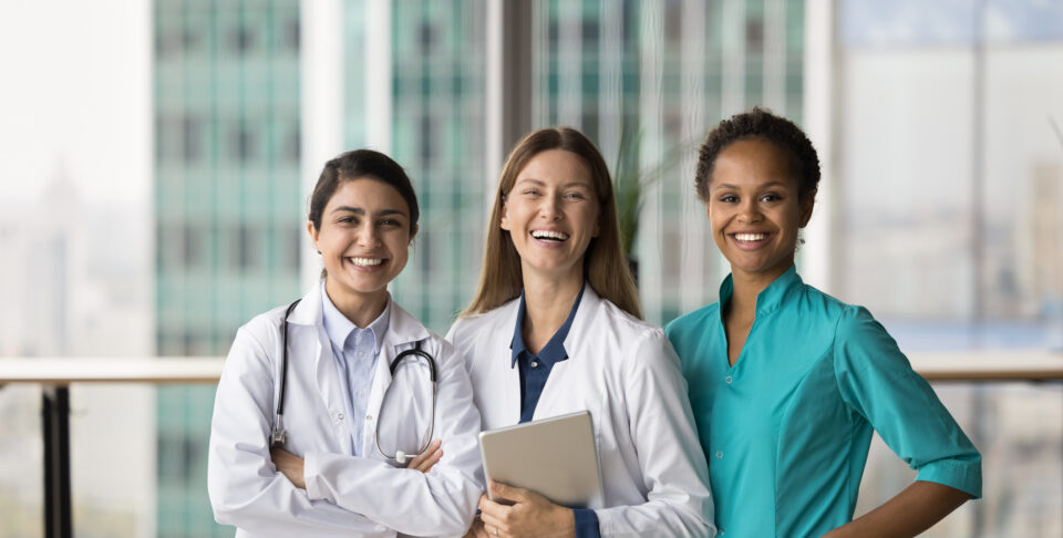 Cheerful diverse medical team of doctors standing together, looking at camera with toothy smiles, laughing. Happy young practitioner women in medic uniform coats posing for portrait