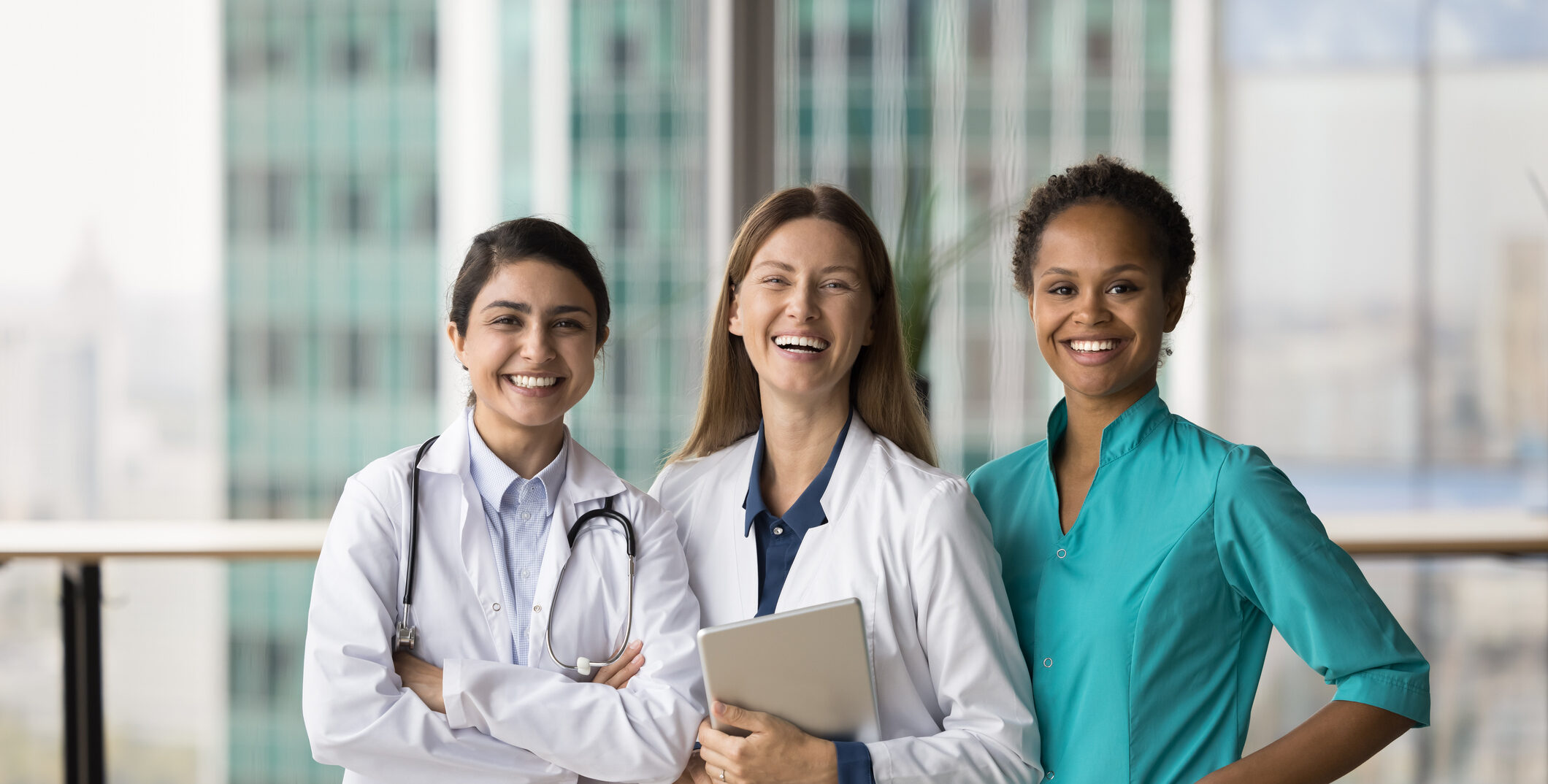 Cheerful diverse medical team of doctors standing together, looking at camera with toothy smiles, laughing. Happy young practitioner women in medic uniform coats posing for portrait