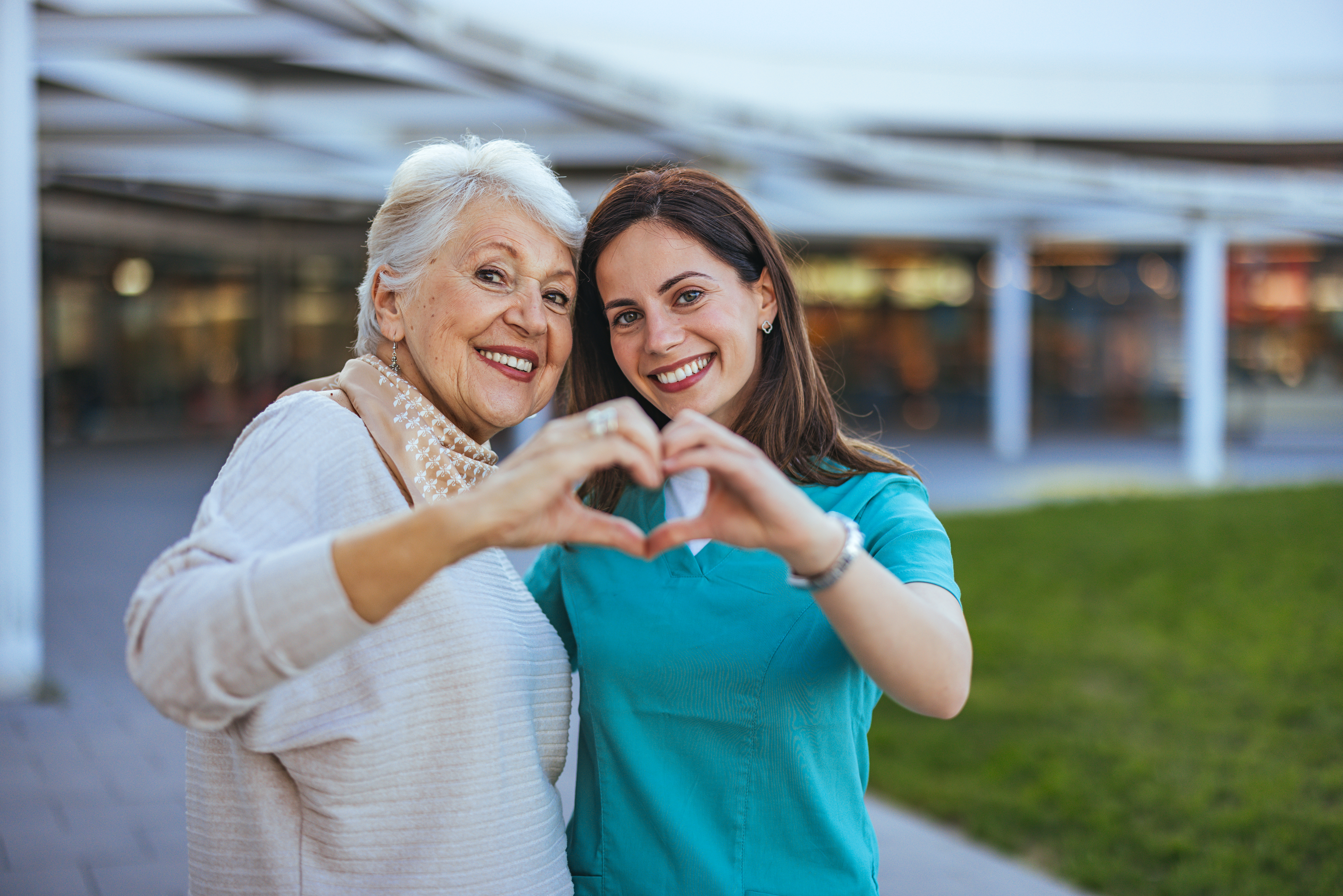 Elderly woman and her caregiver happily form a heart shape with their hands, symbolizing care and companionship in an outdoor setting.
