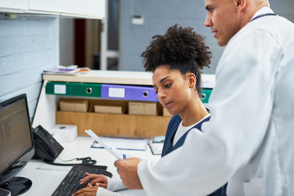 Two healthcare professionals collaborate in a modern office. A uniformed worker refers to documents on a desktop computer while a doctor in a white coat advises. The scene reflects teamwork and efficiency.