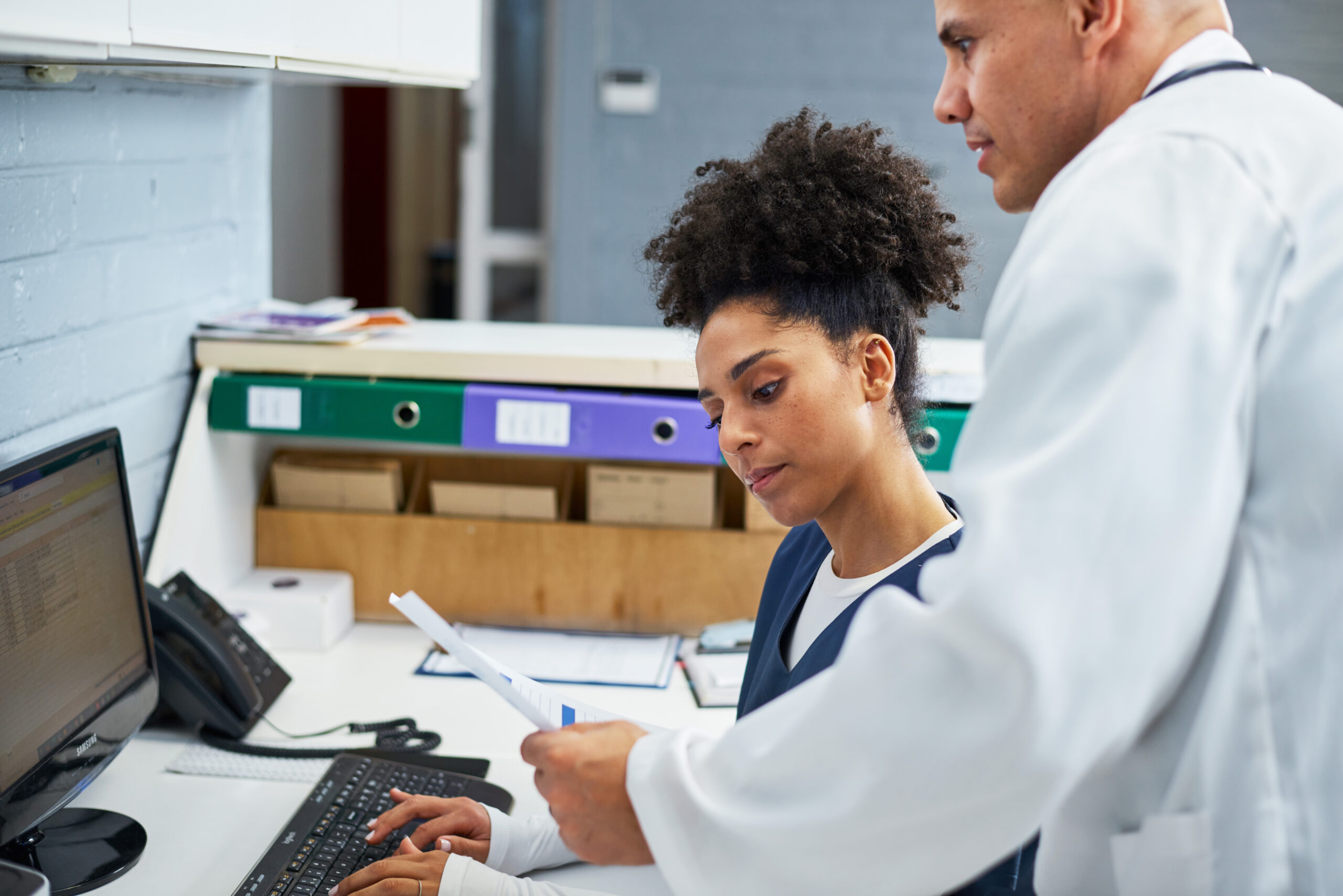 Two healthcare professionals collaborate in a modern office. A uniformed worker refers to documents on a desktop computer while a doctor in a white coat advises. The scene reflects teamwork and efficiency.