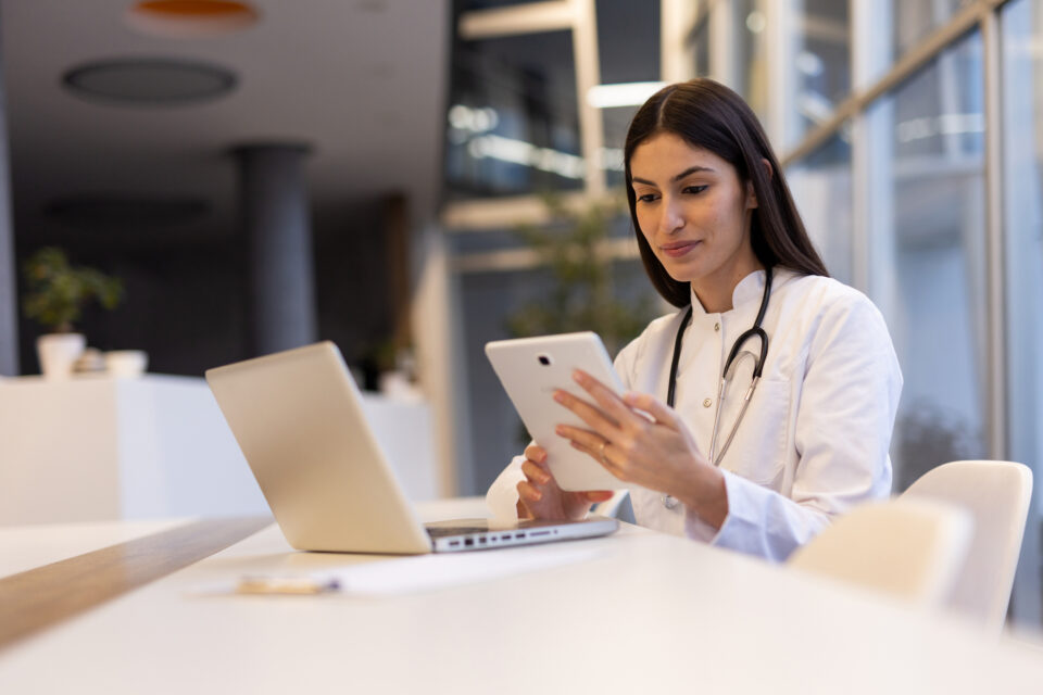 Young female doctor engaging with a digital tablet and laptop in a bright, modern hospital setting, showcasing her expertise in healthcare